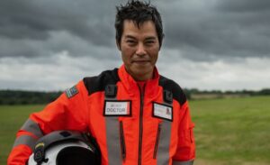 Dr Kevin Fong in an orange air ambulance flight suit, holding a helmet, standing in a field