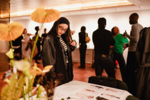 A guest in glasses looks through printed fashion designs on a table while others talk in the background