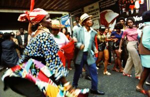 Carnival dancers in the street: a woman in a red headscarf and patchwork skirt and a man in a straw hat and grey jacket dance in front of a crowd