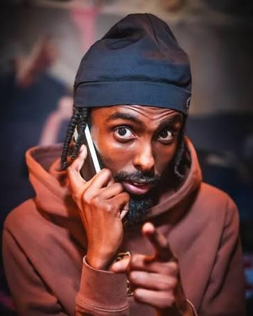 Diesel in a beanie and hoodie, holding a phone to his ear and pointing at the camera