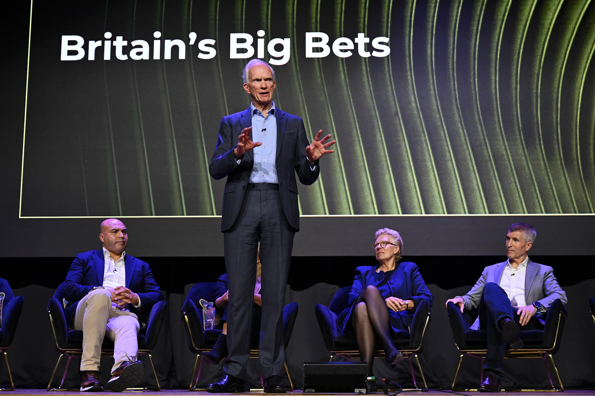 A speaker addresses the room in front of a screen reading Britain’s Big Bets, with a panel seated behind him