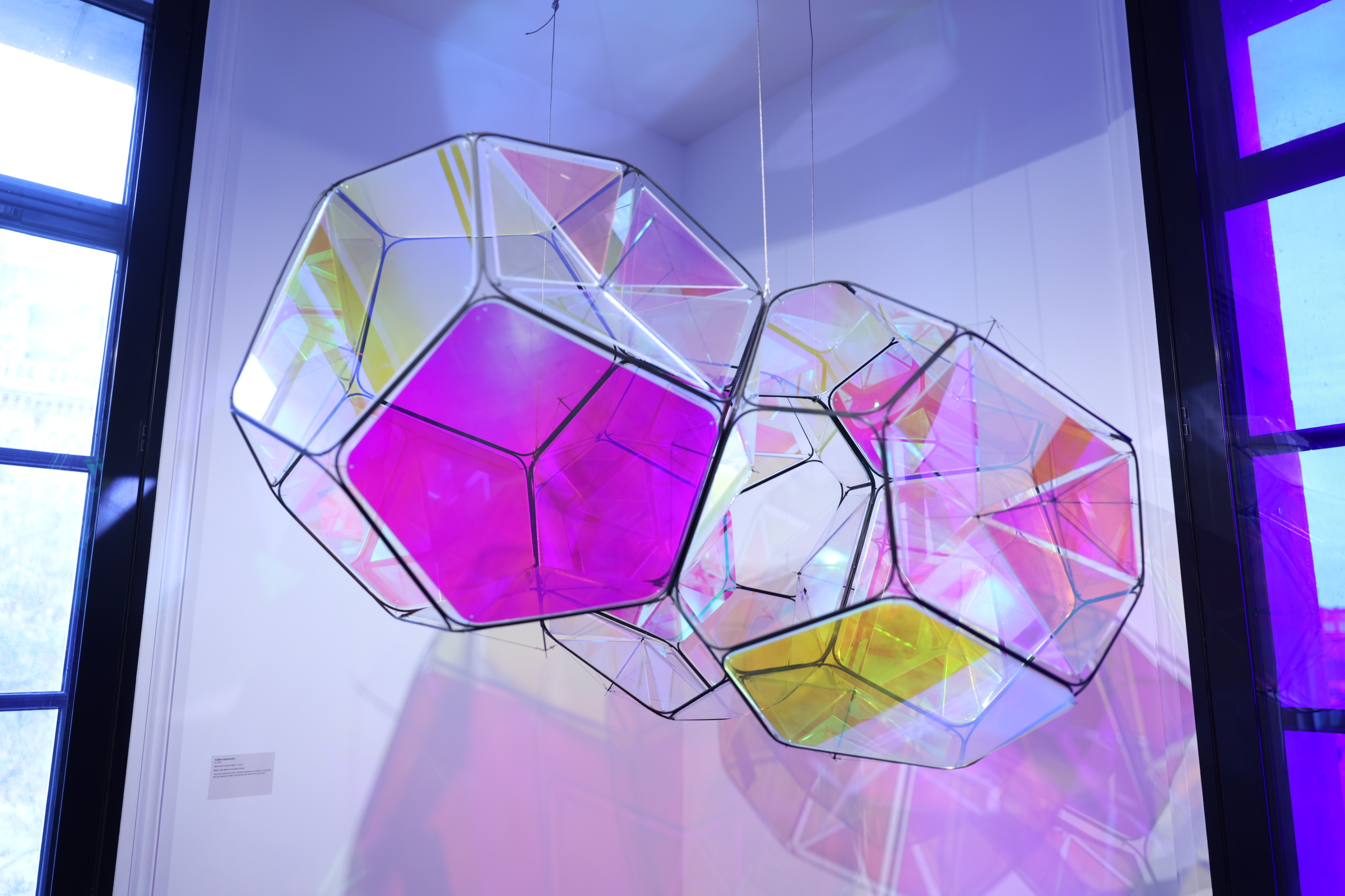 Iridescent polyhedra hang beside a tall window, glowing pink and gold, in The Light Eaters exhibition