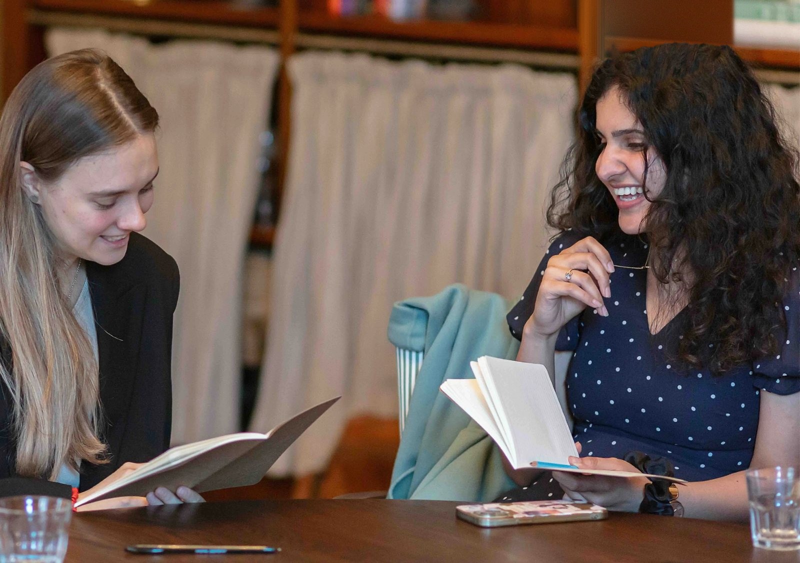 Two women laughing as they read and write in notebooks at a table